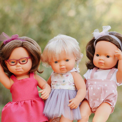 Three Miniland Montessori dolls standing outdoors with blurred trees as a backdrop. The dolls wear colorful summer outfits, each posed with expressive hand gestures and realistic features.