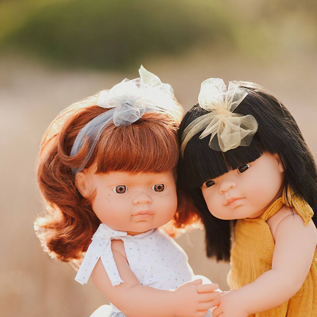 Miniland Montessori red-haired girl doll in a white top standing next to an Asian girl doll in a yellow shirt outside,