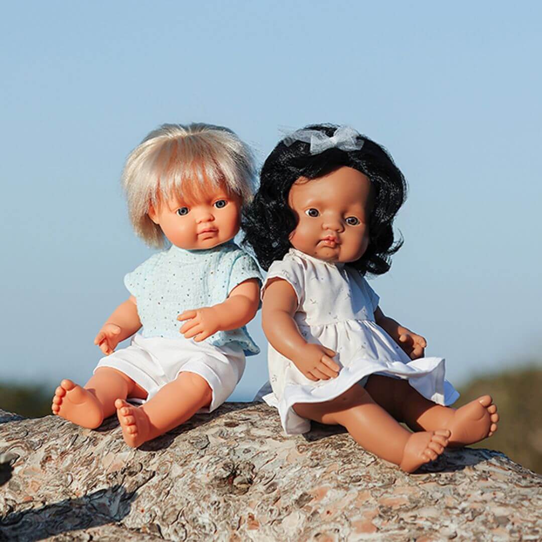 Two Miniland realistic Montessori dolls, one a boy with blonde hair wearing a green dress, and one Hispanic doll with dark hair in a white dress, sitting together on a rock outdoors.
