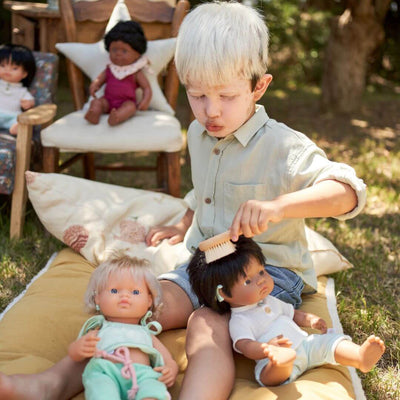 A young child wearing a floral blouse is gently brushing a Miniland doll’s hair using a wooden brush from the Miniland Wooden Doll Beauty Set. The child is seated in on a pillow outside, carefully engaging in nurturing role-play.
