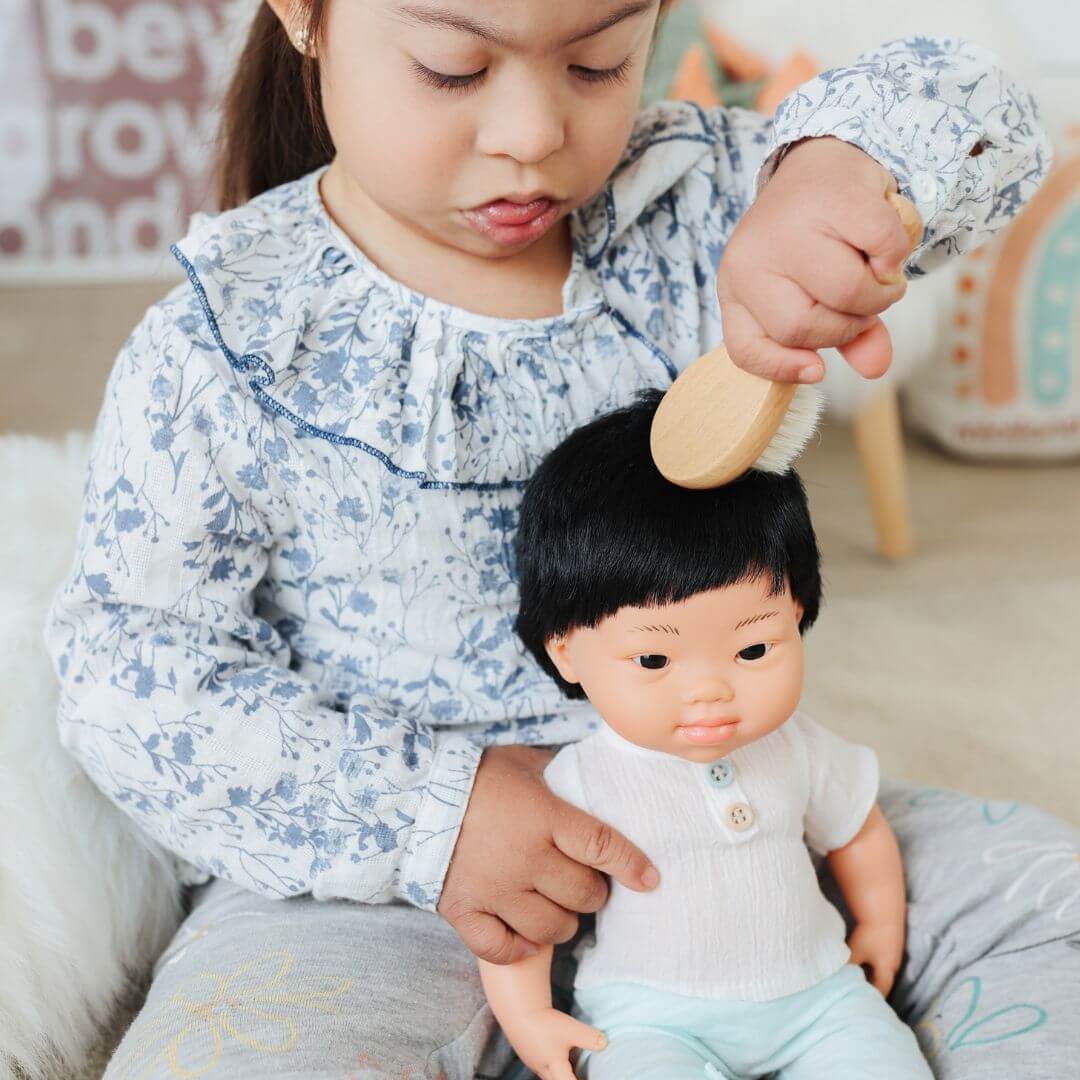 A young child wearing a floral blouse is gently brushing a Miniland doll’s hair using a wooden brush from the Miniland Wooden Doll Beauty Set. The child is seated in a cozy play area, carefully engaging in nurturing role-play.