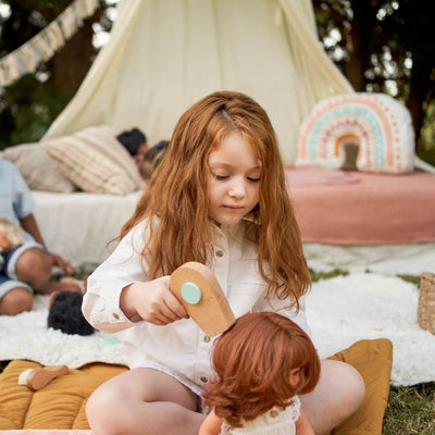 A young girl with long red hair is sitting outdoors on a cozy play mat, using the Miniland Wooden Doll Beauty Set to style a doll’s hair. She is holding a wooden toy hairdryer, while a tent and soft cushions create a whimsical play setting in the background.