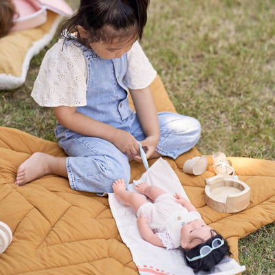 A young child sits on a soft quilted play mat outdoors, using the Miniland Wooden Doll Care Set to change a doll’s diaper on a fabric changing mat. Wooden accessories, including a potty and lotion bottle, are placed nearby, creating a nurturing and imaginative play scene.