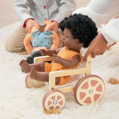 Child pushing a black Miniland doll with curly hair and orange outfit seated in a wooden doll wheelchair, with other dolls and children playing in the background.