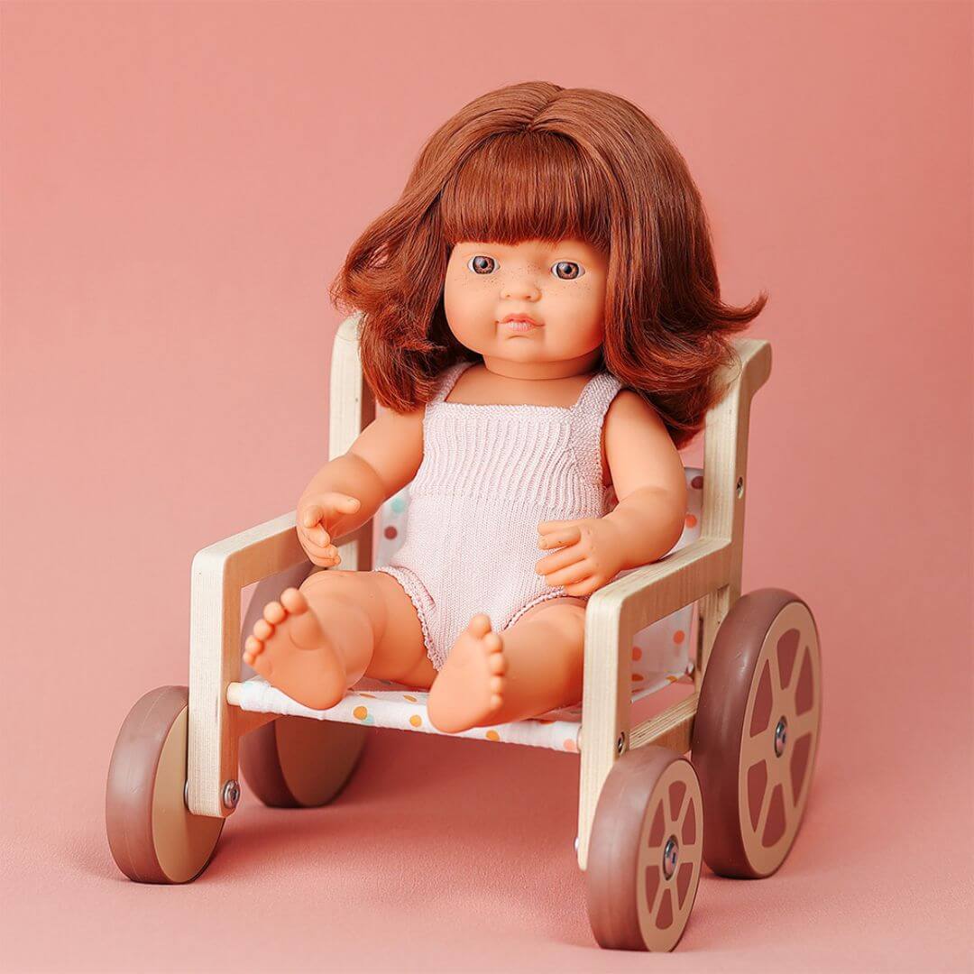 Miniland doll with reddish-brown hair seated in a wooden doll wheelchair with polka dot fabric, shown against a soft pink background.