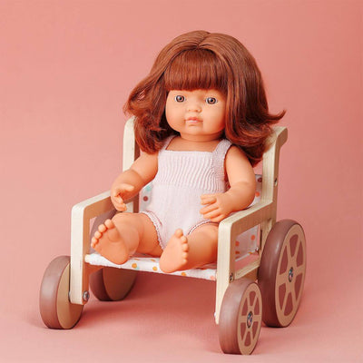 Miniland doll with reddish-brown hair seated in a wooden doll wheelchair with polka dot fabric, shown against a soft pink background.