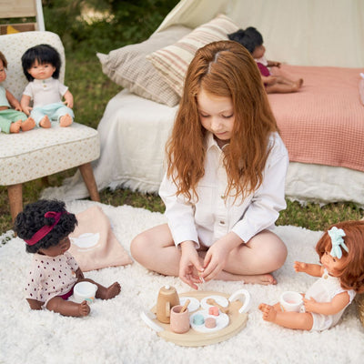 Child setting up the Miniland Wooden Doll Tea Set with teapot, cups, spoons, treats, and napkin arranged on a tray, outside on a blanket for 2 dolls.