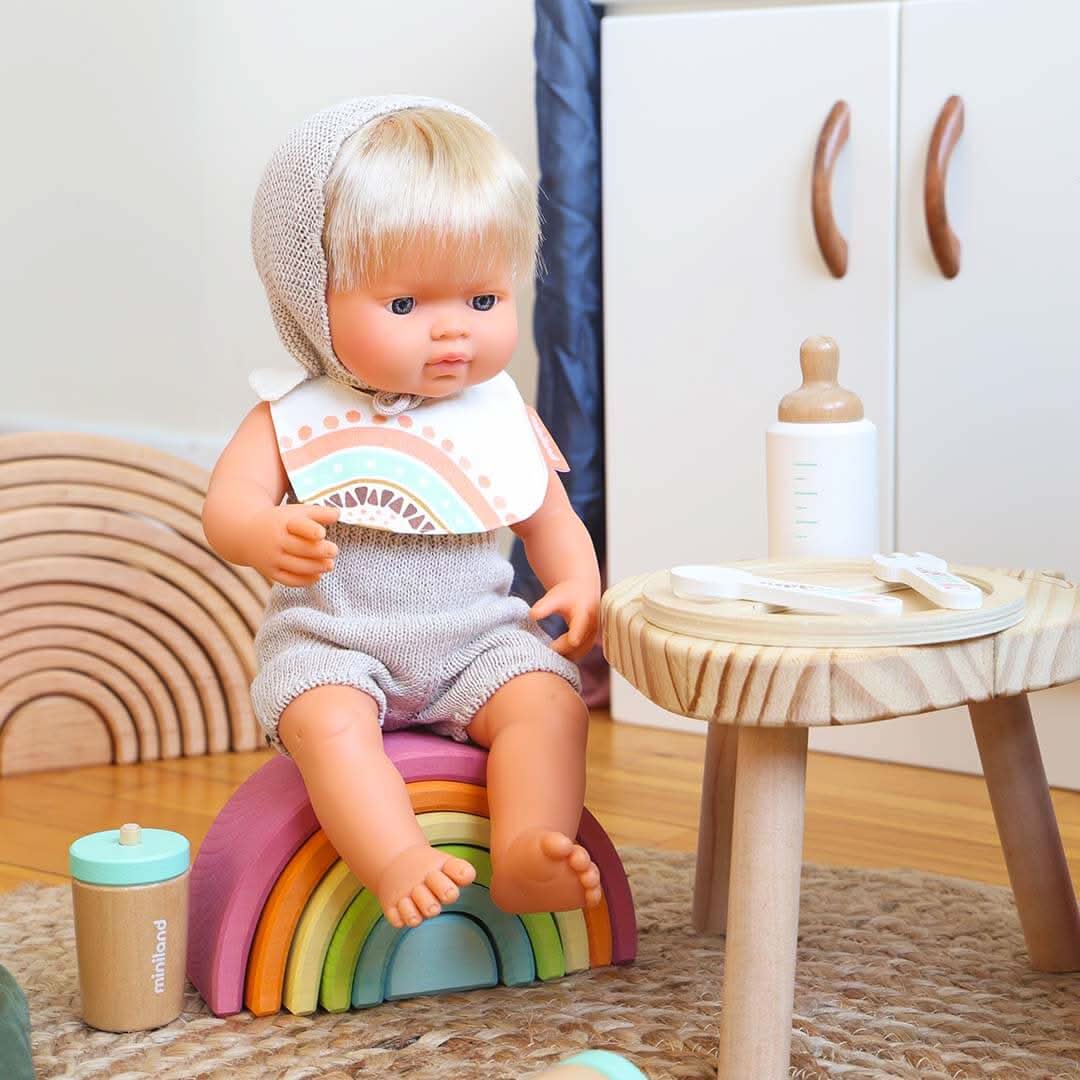 Toy doll with blonde hair sitting on a rainbow-colored toy in a room setting.