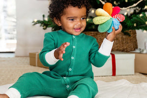 A smiling child in a green onesie holding a HABA Fabric Rainbow Baby Ball, sitting by a decorated Christmas tree.