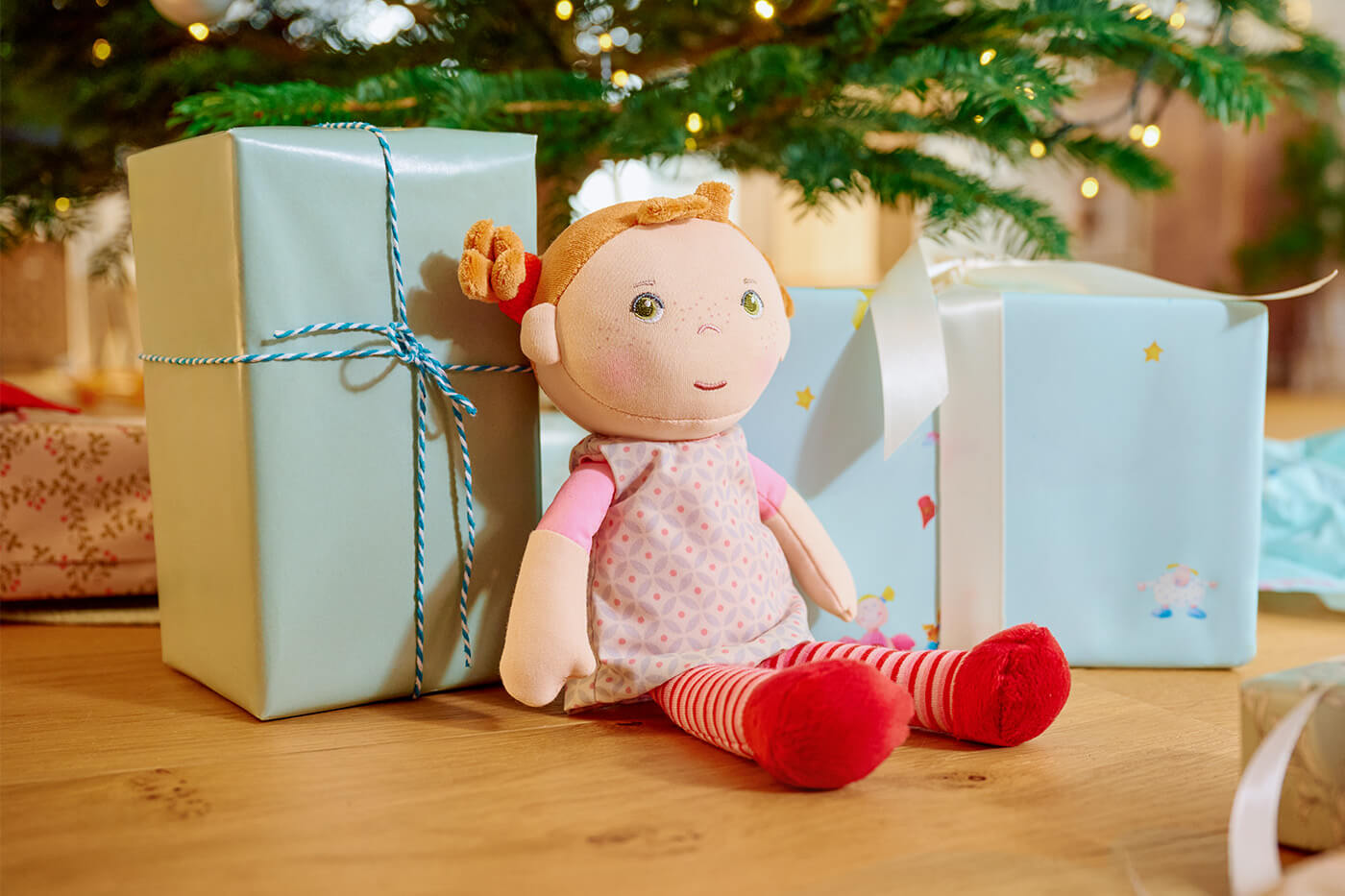A plush Roya from HABA doll sitting next to two wrapped gifts under a Christmas tree.