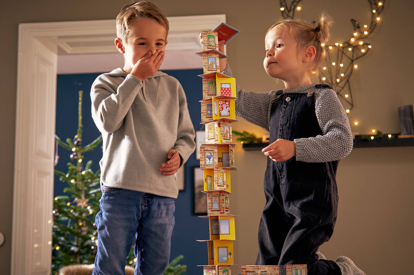 Two children building a tall Rhino Hero card stack next to a decorated Christmas tree indoors.