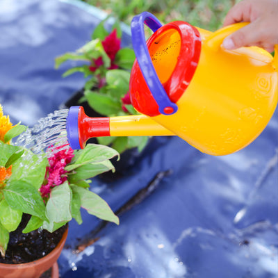 A hand holding a colorful Spielstabil watering can pouring water on plants with green leaves and vibrant flowers.