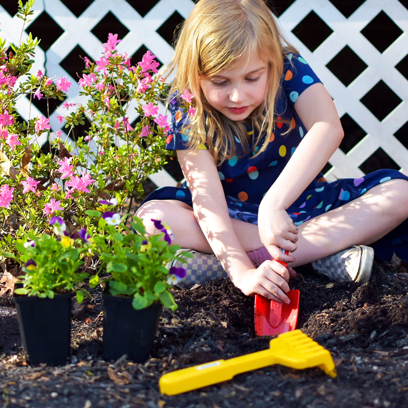 A child in a polka-dot dress digs in the soil with a Spielstabil red shovel, surrounded by flowering plants and a yellow rake.