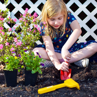 A child in a polka-dot dress digs in the soil with a Spielstabil red shovel, surrounded by flowering plants and a yellow rake.