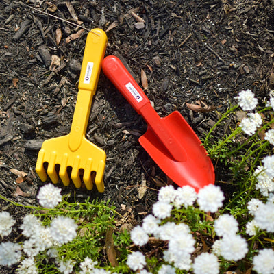 A Spielstabil plastic yellow rake and a red shovel lie on dark soil near white flowers.