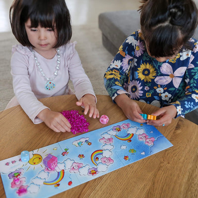 Overhead view of two girls at a table holding game pieces and plastic crystal pieces playing Unicorn Glitterluck Cloud Crystals game